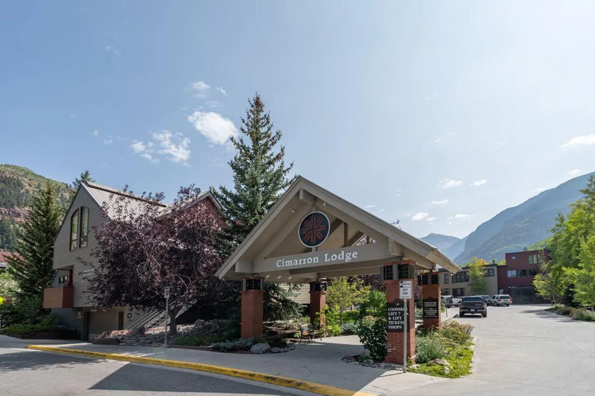 An exterior shot of the Cimarron Lodge sign, surrounded by lush greenery and set against a striking mountain backdrop. The scene suggests easy access to nature and nearby mountain adventures, while the lodge itself represents the starting point for exploring Telluride on foot.