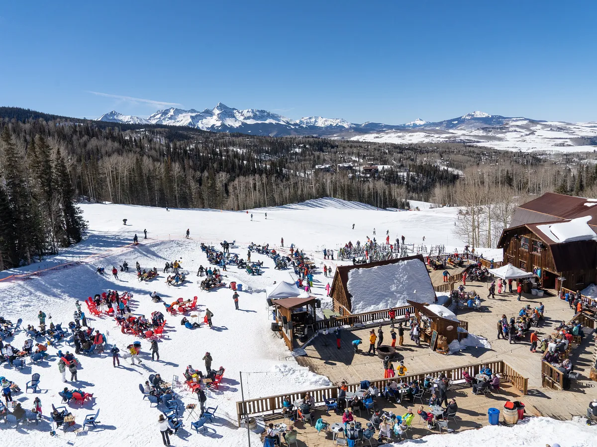 A vibrant scene with people skiing and snowboarding on snow-covered slopes directly in front of the lodge, plus an outdoor seating area and the ski lodge itself in the background. The image exudes the energy and joy of resort living.