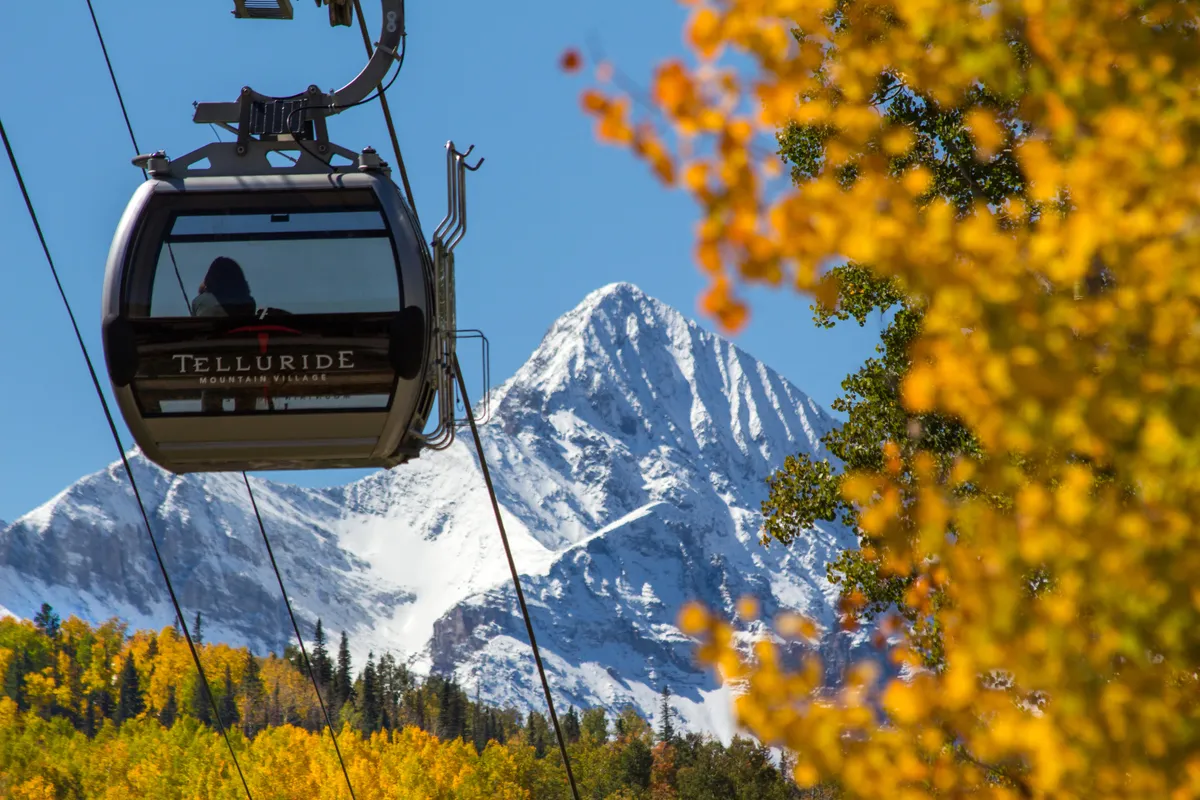 A scenic exterior shot featuring Telluride’s iconic gondola gliding over autumn trees, with snow-capped peaks in the distance. The image beautifully illustrates the town’s easy access to outdoor activities and the unique experience of taking the free gondola up to Mountain Village.