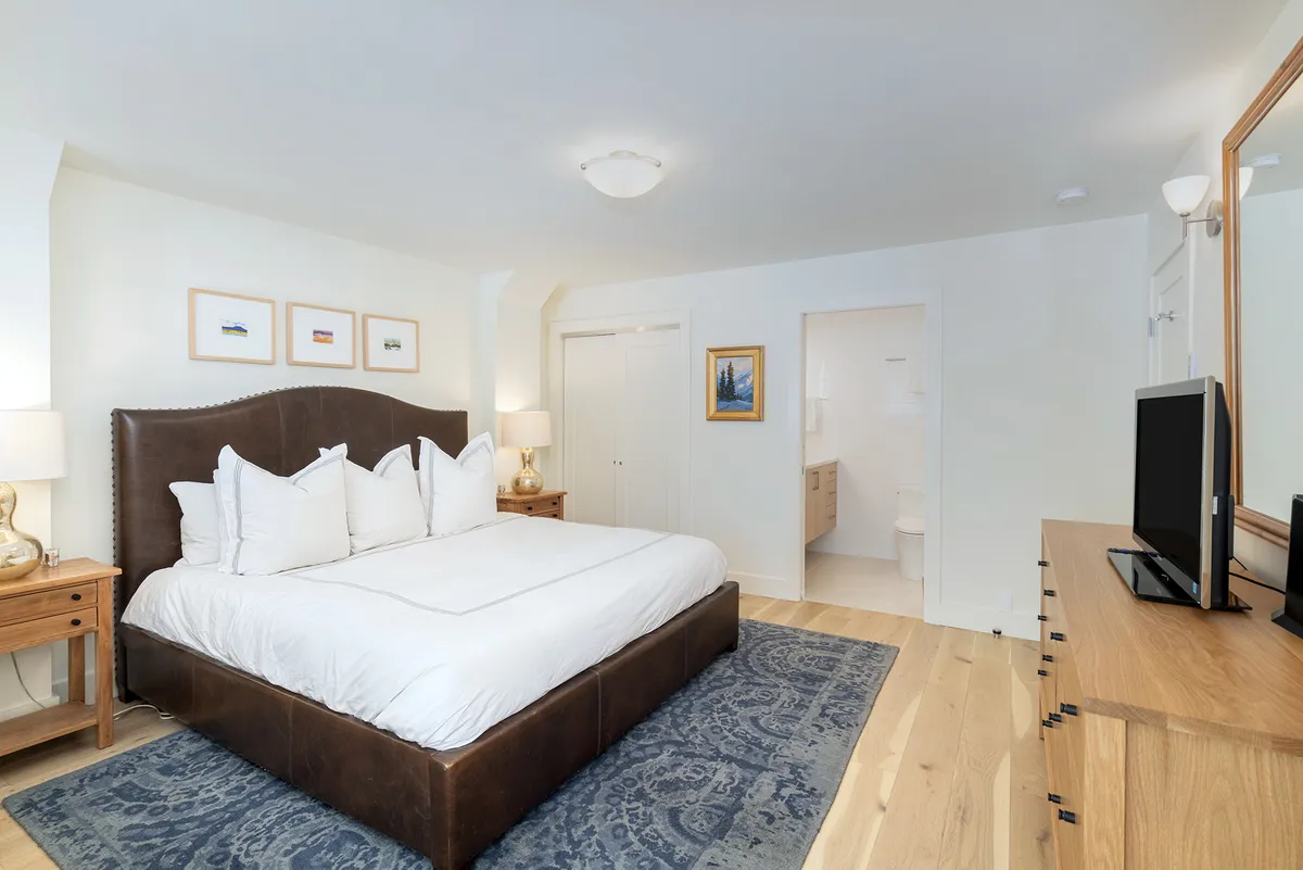 A serene bedroom scene with a large bed featuring plush pillows, matching wooden nightstands, stylish decorative artwork above the headboard, and a television on a wooden dresser. The atmosphere is peaceful and designed for a restful night's sleep.