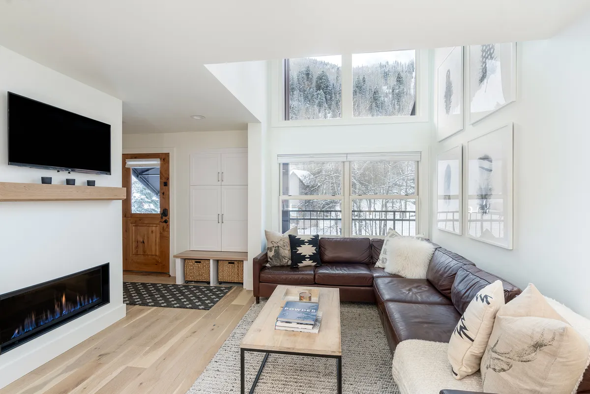 The image shows a spacious and inviting living area with a plush leather sofa positioned beside a cozy fireplace. Floor-to-ceiling windows flood the room with natural light and offer breathtaking mountain views, while a modern coffee table anchors the space. The mood is warm and welcoming, perfect for relaxing after a day on the slopes or gathering with family and friends.