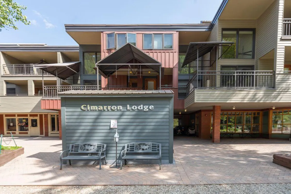 The exterior of Cimarron Lodge with its welcoming sign, benches, and visible balconies, set under gentle light. The scene feels inviting and community-oriented, suggesting easy access to shared amenities and the convenience of being at the heart of the resort area.