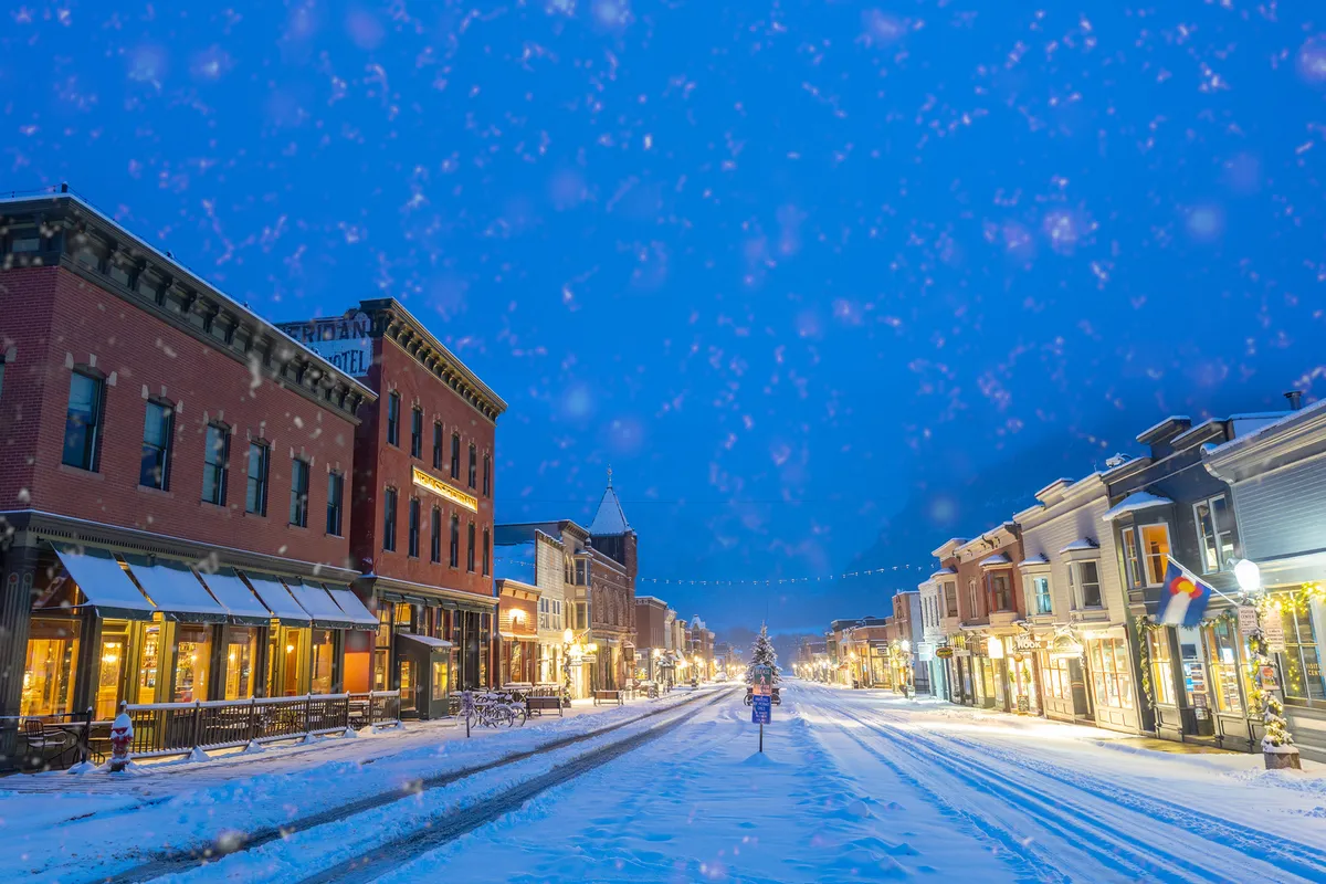 A festive, snow-dusted street scene in Telluride, featuring decorated shops and a distant Christmas tree. The ambiance is warm and inviting, perfect for post-adventure strolls, shopping, and family-friendly seasonal fun.
