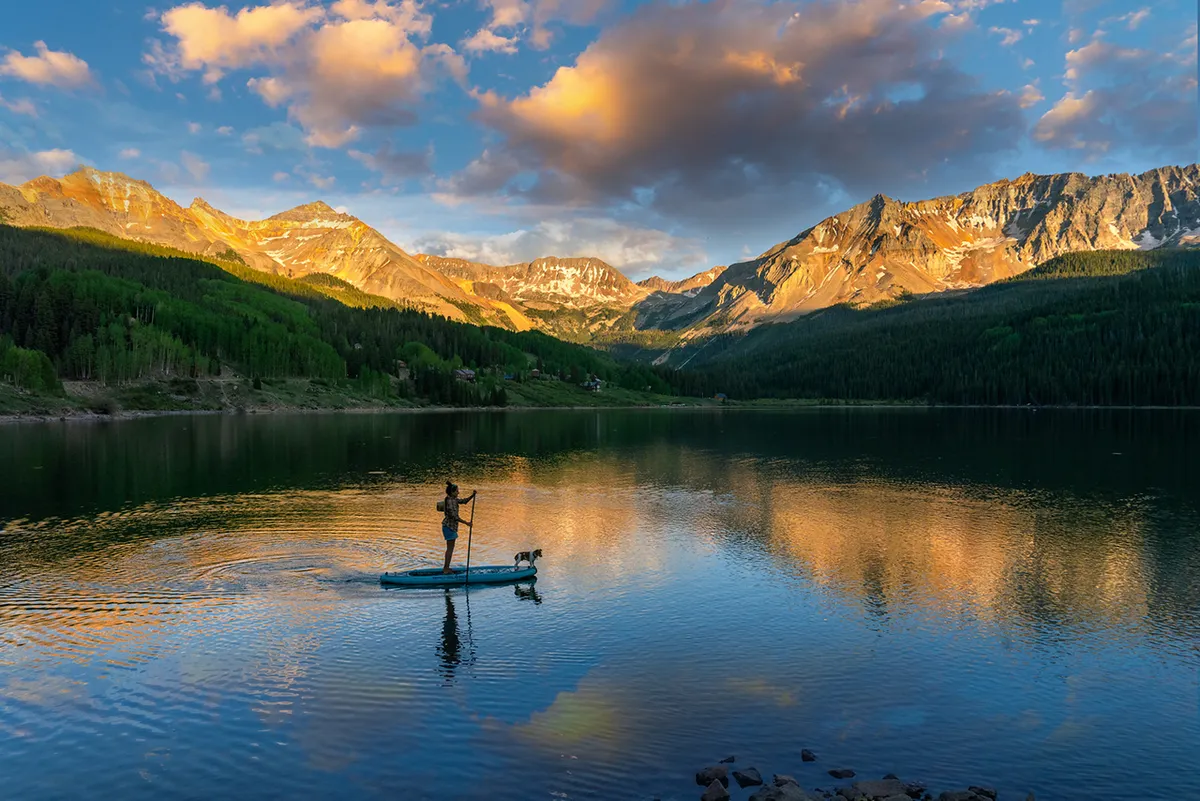 A paddleboarder glides across a serene body of water with their dog, framed by mountains in the background and the soft glow of sunset reflecting on the water's surface. The mood is peaceful and adventurous, highlighting the joy of outdoor recreation in Telluride.