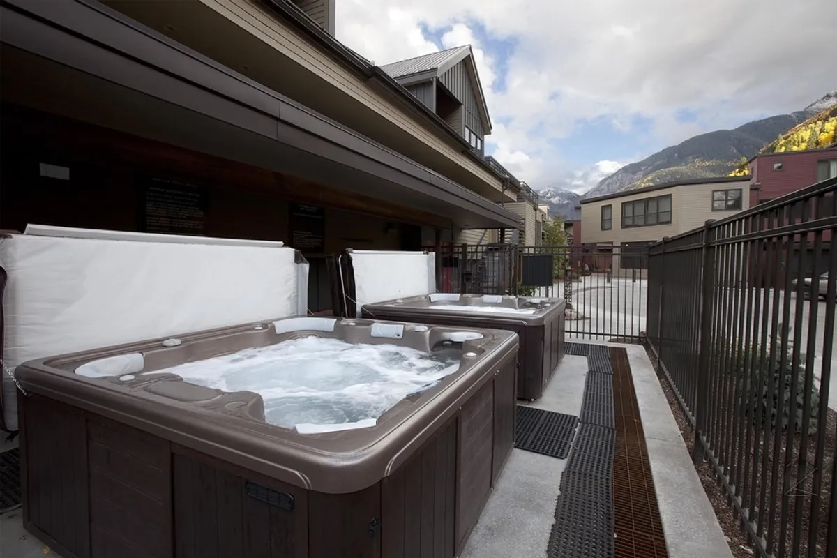 A pair of outdoor hot tubs set against a stunning mountain backdrop, enclosed by a wooden fence for privacy. The tubs are steaming, inviting guests to unwind while soaking in panoramic views after a day on the slopes.