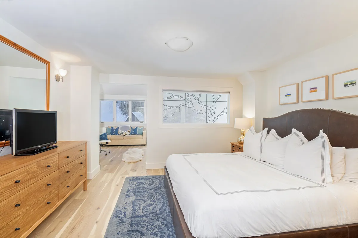 This image features a spacious bedroom with a large bed adorned with decorative pillows, a wooden dresser, a window letting in natural light, and a sofa in the background. The setup suggests an inviting master suite designed for both relaxation and comfort, offering ample space for couples or families.