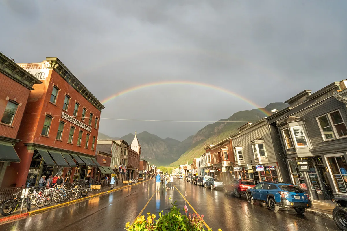 A picturesque view of Telluride’s historic main street with charming brick buildings, a rainbow arching above after rain, and majestic mountains in the background. The wet street and scattered pedestrians evoke a sense of strollable, small-town vibrancy and seasonal beauty.