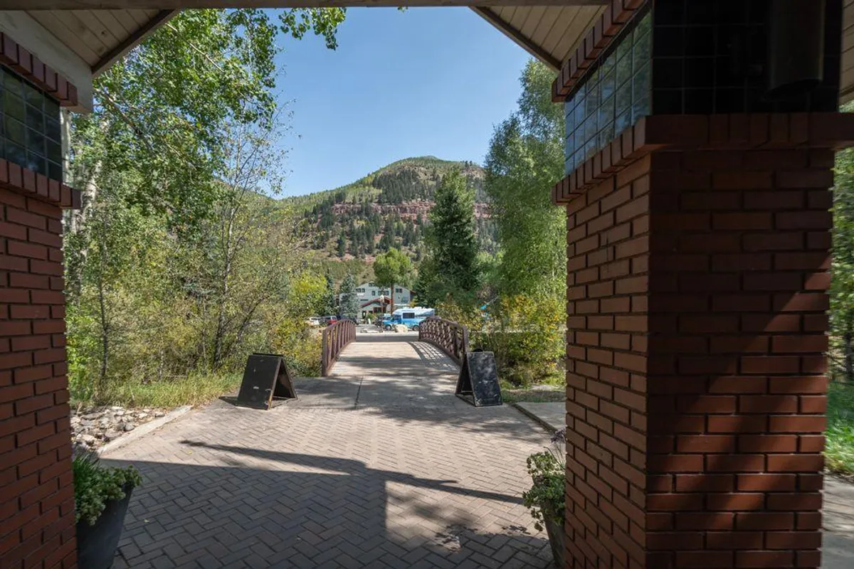 The exterior of Cimarron Lodge features an arched entrance, a welcoming pathway, a picturesque bridge, and is surrounded by lush trees and a dramatic mountain backdrop. The scene suggests easy access to outdoor adventures and a harmonious connection to nature.