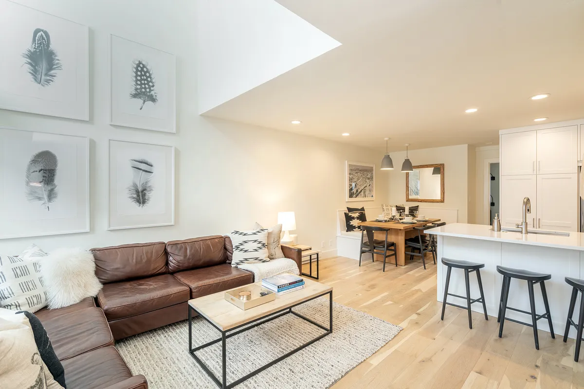 This image features an inviting great room with plush leather sofas, a dining table set for six, and eye-catching artwork on the walls. The open-concept layout seamlessly connects the living area to a well-appointed kitchen island, creating a sense of togetherness. Sunlight streams in through generous windows, bathing the space in natural light and highlighting its cozy, sociable mood.